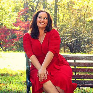 Picture of Dellany Peace, a white woman with shoulder length brown hair posing on a bench in a park, wearing a bright red dress, hands on her knee, and a whimsy bright smile on her face.