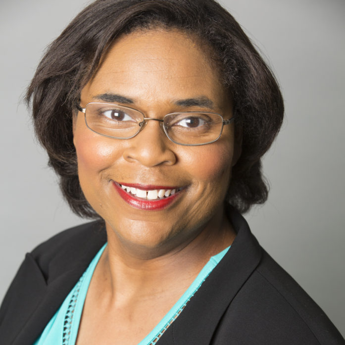 Headshot of Kim Scott, a Black woman posing in front of a neutral background, wearing glasses, a black jacket and blue t-shirt.
