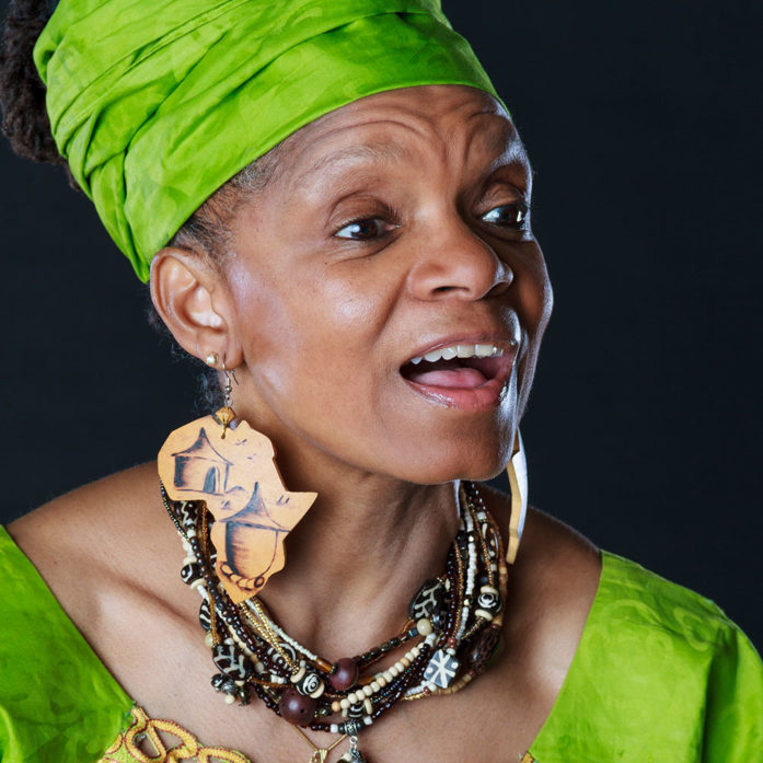 Headshot of Janice Curtis Greene, a Black woman wearing a bright green top and a head wrap in the same color, big earrings in the shape of the African continent, and a big beaded necklace.