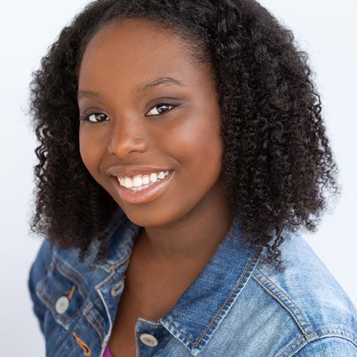 Headshot of Anah Ambuchi, a young Black girl smiling brightly, posing in front of a white backdrop, wearing a denim jacket, and her hair down to her shoulders.