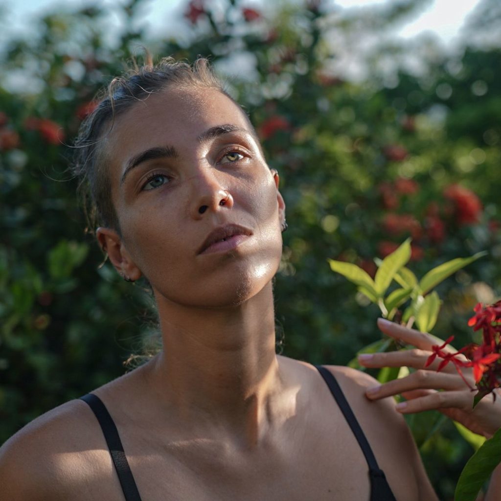 Headshot of Manon de Reeper, a Black woman posing artfully among flowery bushes, wearing a black top with thin straps.