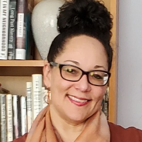 Headshot of Carrie Curtis Young, a Black woman posing in front of a bookcase with a smile on her face, her hair in an up-do, wearing glasses and gold earrings, a tan scarf and orange top.