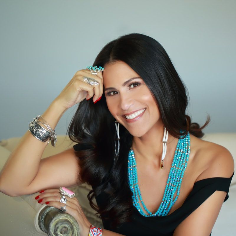 Headshot of Dr. Joely Proudfit, an American Indian woman posing on a chair leaning her head against a hand. She has long dark hair and a bright smile, wearing a black dress and a turquoise beaded necklace, and long beaded earrings, and many silver rings and bracelets..
