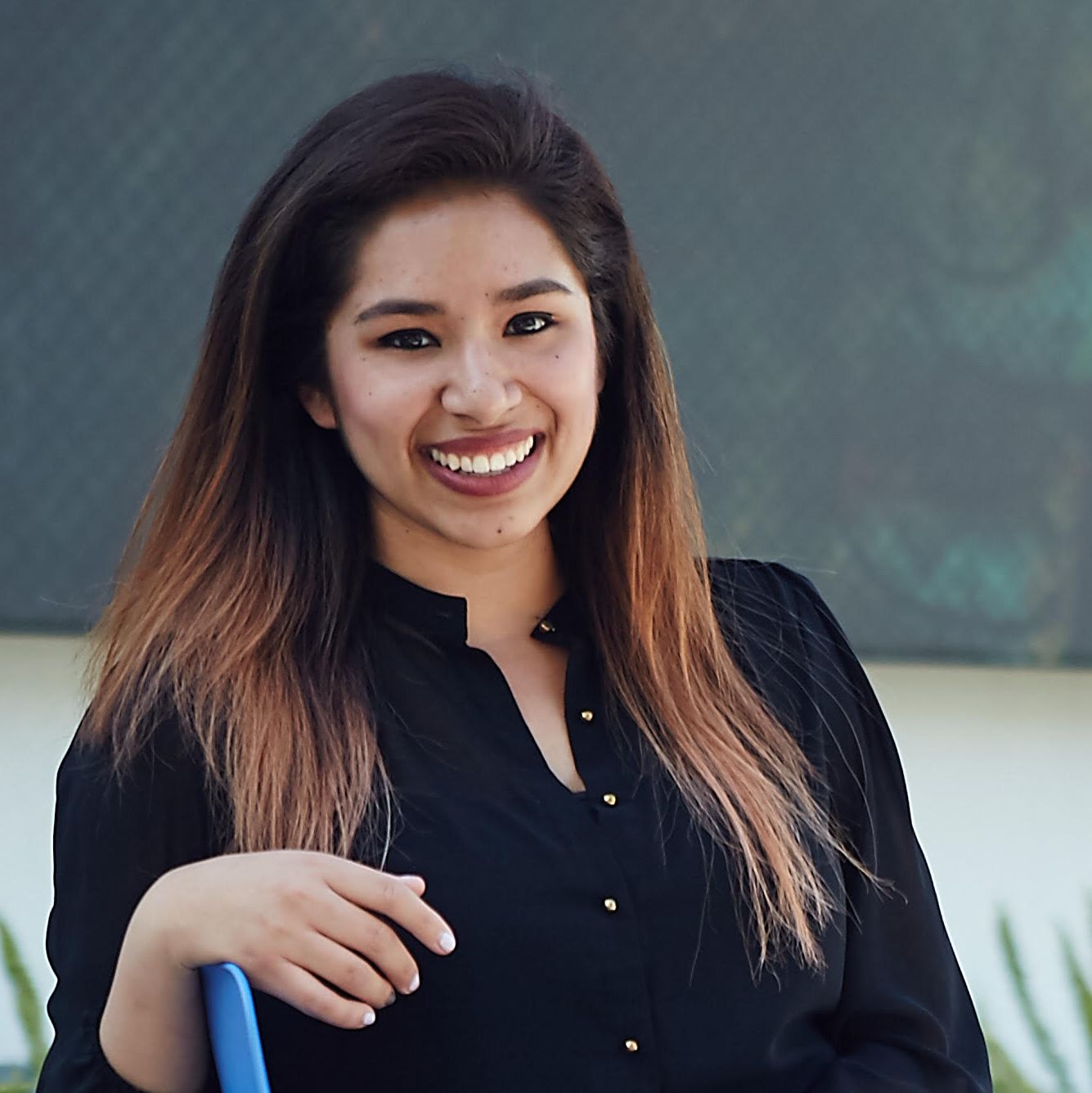 Headshot of Karissa Valencia, an American Indian woman smiling brightly, casually posing in front of a dark grey backdrop, wearing a black shirt and her long hair down.