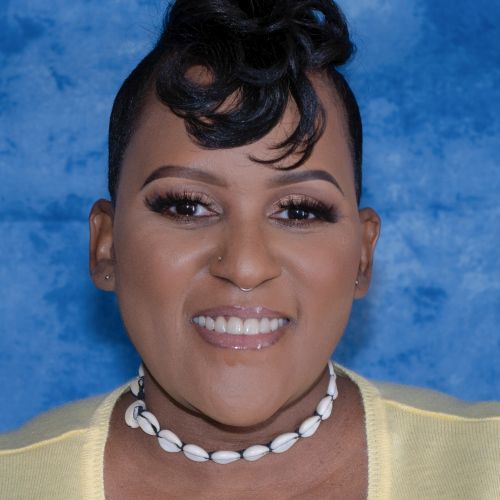 Headshot of Kathryn Washington, a Black woman posing in front of a blue backdrop, wearing her hair in an up-do, a yellow shirt and a shell necklace, and a bright smile.