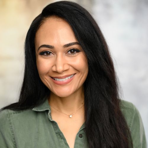 Headshot of Charlita Gaston, a Latina woman posing in front of a neutral background, with long dark hair, wearing a moss green shirt and smiling brightly.
